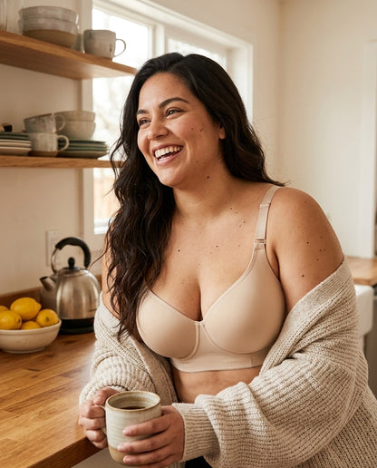 A woman laughing in her kitchen with a coffee, wearing The Ease bra in Beige under a cardigan